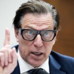 A man in a suit and glasses speaks emphatically, raising his index finger during what appears to be a formal hearing or meeting.