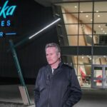 A man wearing a dark jacket stands outside the Alaska Airlines Center at night, with the buildings illuminated sign and glass entrance visible behind him.
