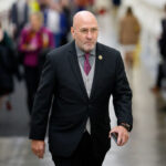 Rep. Clay Higgins in a suit and red tie walks indoors, holding a phone. People are blurred in the background, and the setting appears to be a hallway or corridor with white walls and a shiny floor.