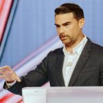 A man in a suit speaks at a podium, gesturing with his right hand as he addresses the crowd about the challenges posed by infidels. The background features geometric red and blue designs.
