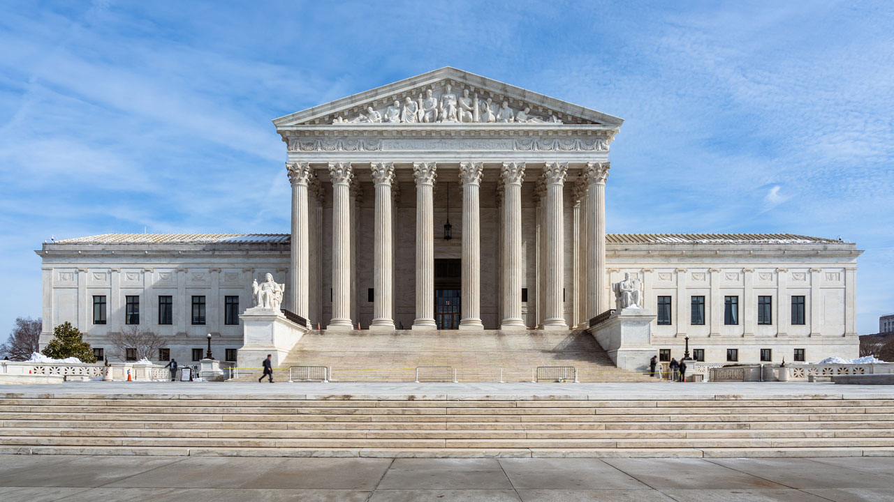 The United States Supreme Court building with tall columns, a triangular pediment, statues, and broad stone steps under a blue sky.
