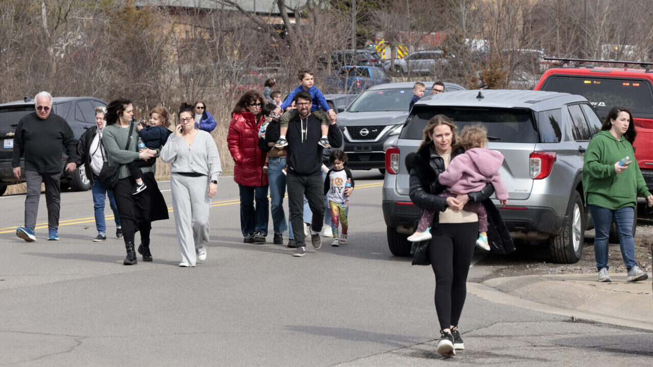 Home 1 A group of adults and children walk together on a street near parked cars. One woman in front carries a child. Trees and emergency vehicles are in the background.