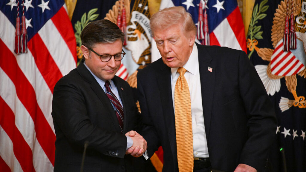 Kevin Roberts Makes the Case for Keeping Conservatives in Power 1 Two men in suits shake hands in front of multiple U.S. flags and official seals. One man wears glasses, and the other has blonde hair and an orange tie. They appear to be at a formal event or ceremony.