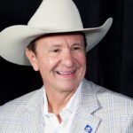 A smiling man in a white cowboy hat and a light plaid blazer stands in front of a dark background.