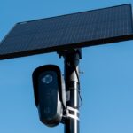 A black security camera is mounted on a pole beneath a large rectangular solar panel against a clear blue sky.