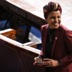 Ilhan Omar in a maroon blazer and patterned headwrap smiles while standing in a large, formal room with wooden seats and a round table, holding a phone.