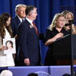 A woman speaks at a podium with the presidential seal, flanked by others. A man stands beside her and a young woman holds a photo of a child. President Donald Trump and woman are in the background.