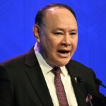 A man in a suit and tie speaks into microphones, gesturing with his right hand, against a blue background.