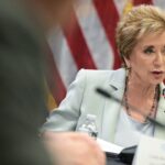 A woman in a light suit speaks into a microphone at a meeting focused on education, with an American flag in the background and a nameplate visible in front of her.