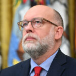 Russell Vought with glasses, a gray beard, and a suit with a red tie looks upward. Colorful flags and a blurred emblem are visible in the background. In the Oval Office.
