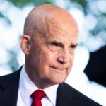 Keith Self in a dark suit, white shirt, and red tie, standing outdoors with blurred green foliage in the background.