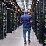 A person wearing a blue shirt, jeans, and a white cap walks down an aisle between tall server racks in a brightly lit data center.