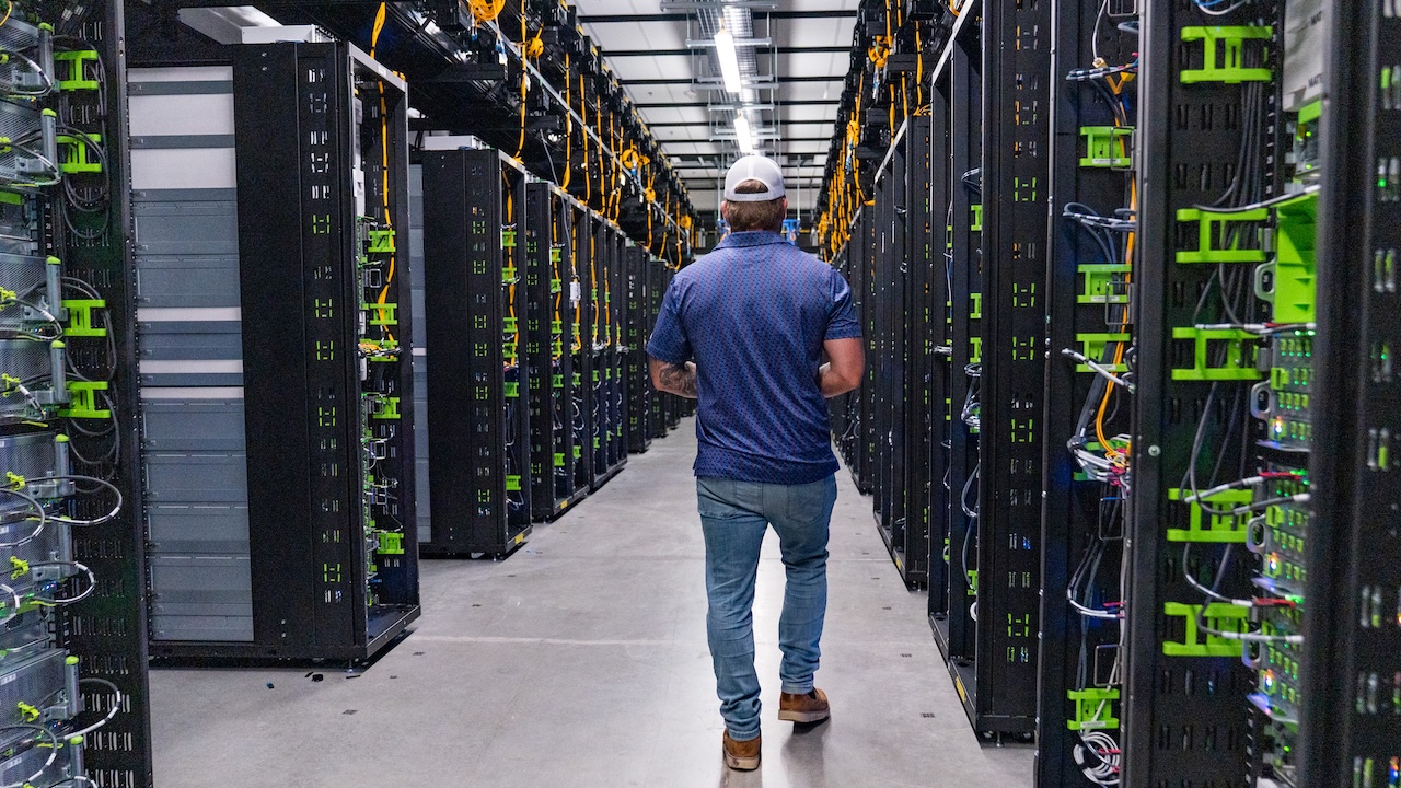 A person wearing a blue shirt, jeans, and a white cap walks down an aisle between tall server racks in a brightly lit data center.