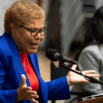 An older woman in a blue blazer speaks passionately into a microphone, gesturing with her hands. Another woman sits beside her, listening and smiling.