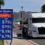 Exxon gas station sign displays fuel prices; regular at $5.79, extra at $5.99, supreme at $6.19, and diesel at $6.79 per gallon. A white semi truck drives by on a sunny day.
