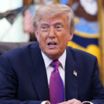 President Trump in a navy suit and purple tie sits at a desk, speaking. Flags and decorative items are visible in the blurred background.