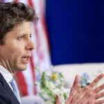A man in a suit speaks and gestures with his hands, perhaps discussing the impact of AI. There is an American flag and some flowers in the background.