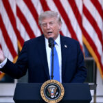 President Donald Trump in a suit speaks at a podium with the U.S. presidential seal, in front of several large American flags.