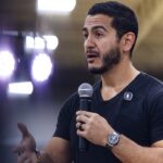 A man with dark hair and beard speaks into a microphone, wearing a black shirt and watch, gesturing with his left hand. Bright lights and a blurred background suggest he is at an indoor event or conference.