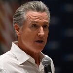 A man with gray hair wearing a white shirt speaks into a microphone. An out-of-focus American flag is visible in the background.
