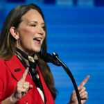 A woman in a red blazer speaks energetically at a microphone against a blue background, gesturing with both index fingers raised.