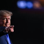 Republican President Donald Trump points forward with a serious expression, standing against a dark background with blurred lights in the upper right corner.