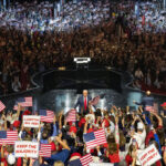 A large crowd waves American flags and Keep the Majority signs at an indoor political rally, with President Donald Trump speaking on stage in the center background.