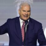 A man in a navy suit and striped tie speaks passionately at a podium, gesturing with his right hand. A blurred background with white star shapes is visible behind him.