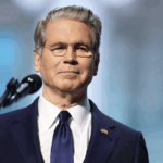 Scott Bessent in a navy suit and tie stands in front of a microphone, looking forward with a neutral expression. He has gray hair, glasses, and an American flag pin on his lapel. The background is blurred.