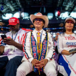 Four people sit at a political rally wearing red, white, and blue pro-Trump apparel, holding signs that read BIGGER PAYCHECKS FOR WORKING FAMILIES. The crowd is visible in the background, with fellow Republicans showing their support.