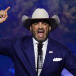 Webb County Judge Tano Tijerina in a blue suit and cowboy hat speaks passionately at a podium, raising his right hand with his index finger pointed upward. An American flag pin is on his lapel, and the background features a blue, starry pattern.