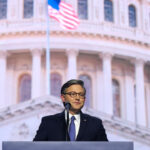 Speaker Mike Johnson in a suit and glasses stands at a podium with a microphone, speaking in front of a large image of the U.S. Capitol dome. An American flag is visible in the background.