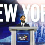 A man in a suit stands at a podium labeled Dallas with a cityscape and the word NEW YORK displayed on a large screen behind him.
