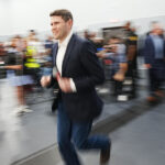 A man in a dark blazer and jeans jogs indoors with a blurred background of people and cameras, giving a sense of motion and energy.
