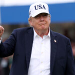 President Donald Trump, dressed in a dark blazer and white USA cap, raises his fist while standing outdoors, looking ahead, with blurred people and a blue canopy in the background.