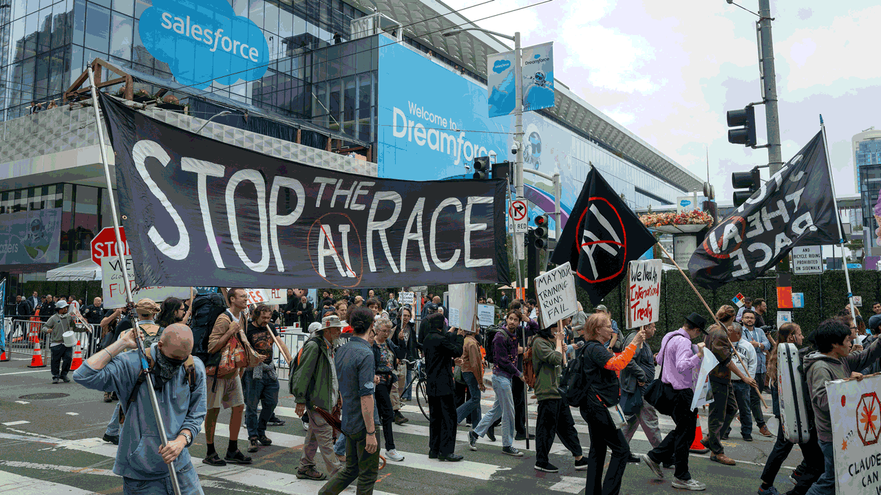 Protesters cross a city street holding signs, including a large banner reading Stop the AI Race, in front of a modern building with Salesforce and Dreamforce banners.