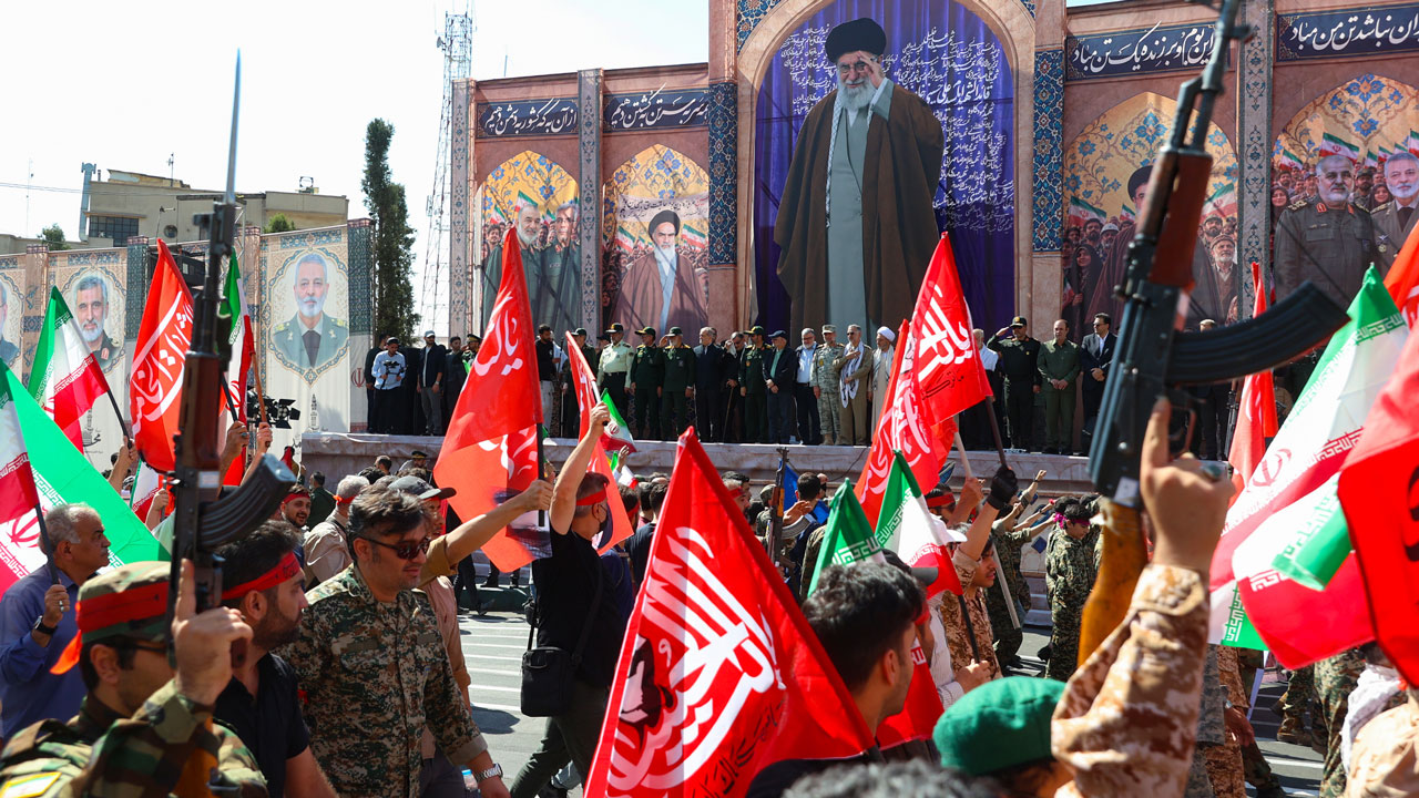 A crowd of people holds red and green flags during an Iran rally in front of large murals depicting political and military leaders.