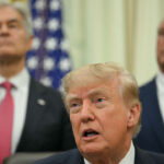 Three men in suits stand indoors; the man in front is speaking, while two others stand behind him. U.S. flags and official emblems are visible in the background.