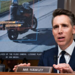 A man in a suit speaks at a hearing, seated at a desk labeled Mr. Hawley. Behind him, a screen displays a vehicle image with highlighted license plate data and analysis from a camera system.