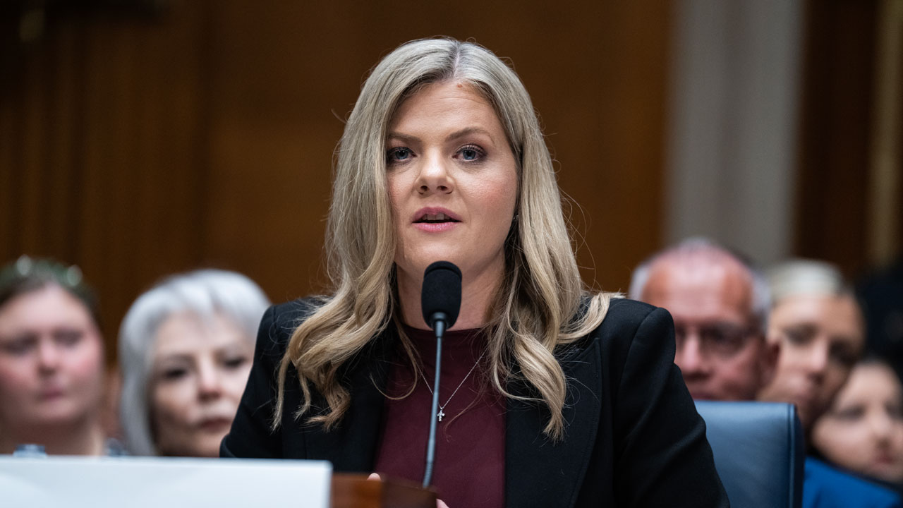 A woman with long blonde hair speaks into a microphone at a formal hearing, wearing a black blazer and maroon top. Several people sit blurred in the background.