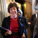 Sen. Susan Collins in a red dress and black blazer, smiling, holds folders while standing near an elevator in a building with marble columns. Other people are around her.