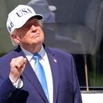 Donald Trump in a hat stands outdoors, raising his fist and smiling.