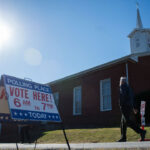 A person walks past a Vote Here! sign outside a brick church in Virginia under a bright sun, indicating the location is a polling place.