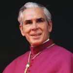 An older man in religious attire with a red robe and gold chain stands at a microphone, smiling slightly, against a dark background.