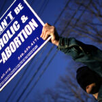 A person holds a large sign reading, You cant be Catholic & pro-abortion, with contact information and a website, against a blue sky and some tree branches.