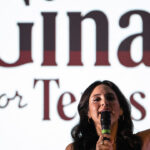 A woman with dark hair speaks into a microphone, raising one finger, in front of a large screen displaying partially visible text that reads Gina and for Texas..
