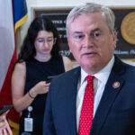 A man in a suit and red tie speaks while three women stand behind him, one holding a phone. Office plaques and flags are visible in the background.