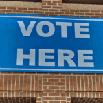 A blue sign with white letters reads VOTE HERE and is attached to a brick wall, indicating a polling location.