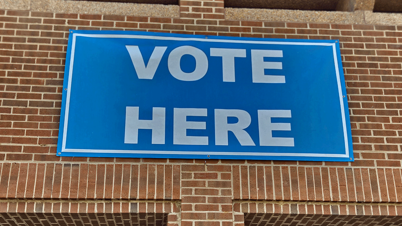 A blue sign with white letters reads VOTE HERE and is attached to a brick wall, indicating a polling location.