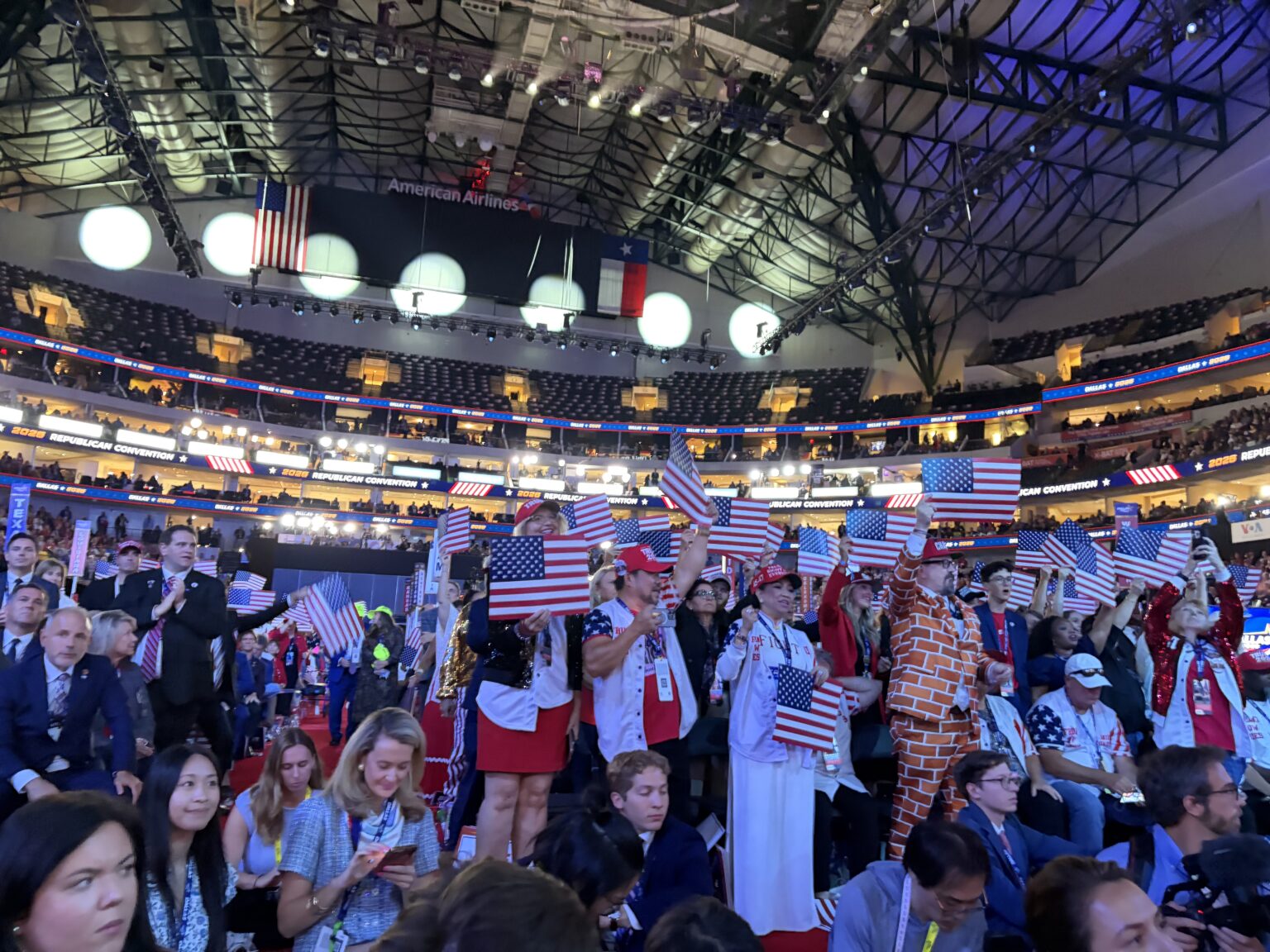 A crowded indoor arena at the Republican National Midterm Convention with people dressed in patriotic outfits, many holding small American flags.